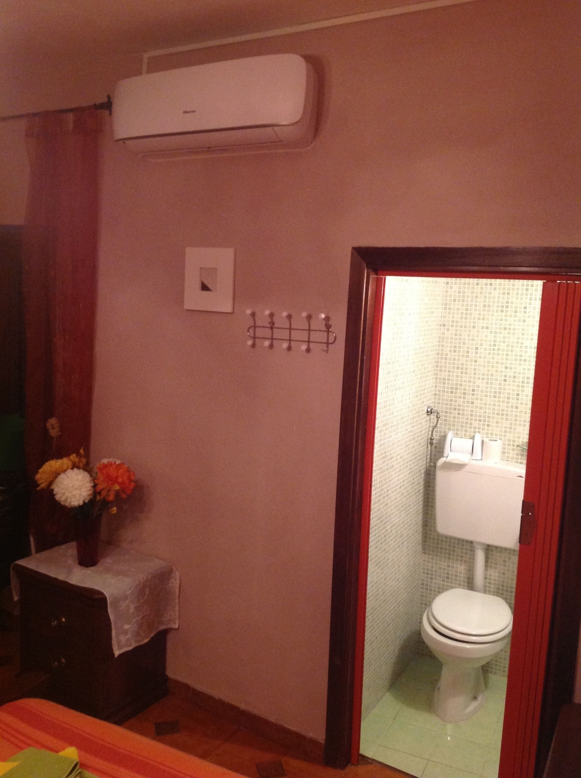 Un baño con inodoro, lavamanos y ducha. Hay un espejo, un perchero para toallas y una mesita con un jarrón de flores.