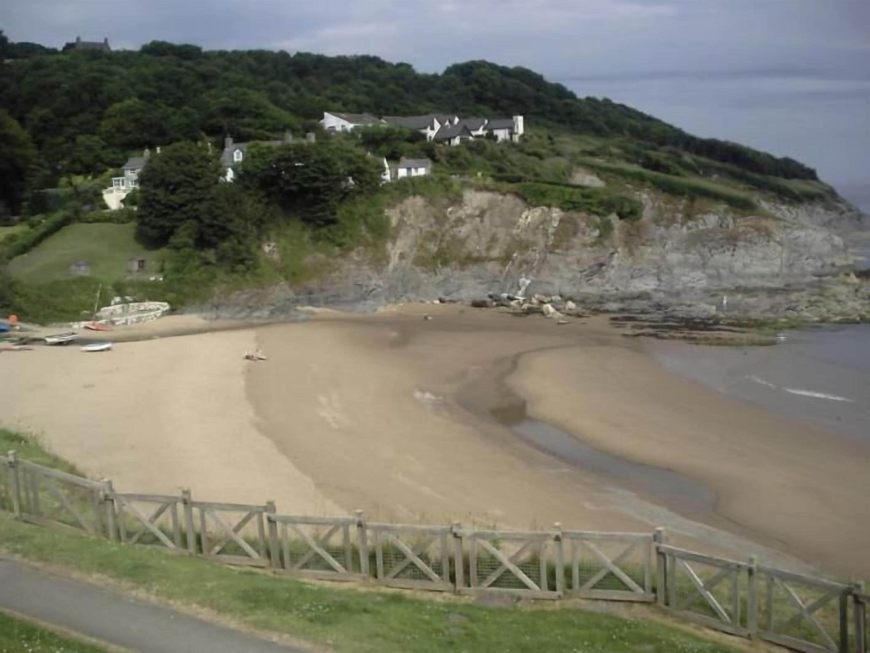 Una spiaggia sabbiosa con una recinzione in legno in primo piano, un litorale roccioso e case su un pendio sullo sfondo.