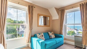 A room with a blue sofa, large windows with curtains, a radiator, and a mirror on the wall. - Rooms at The Highcliffe (Cardigan)