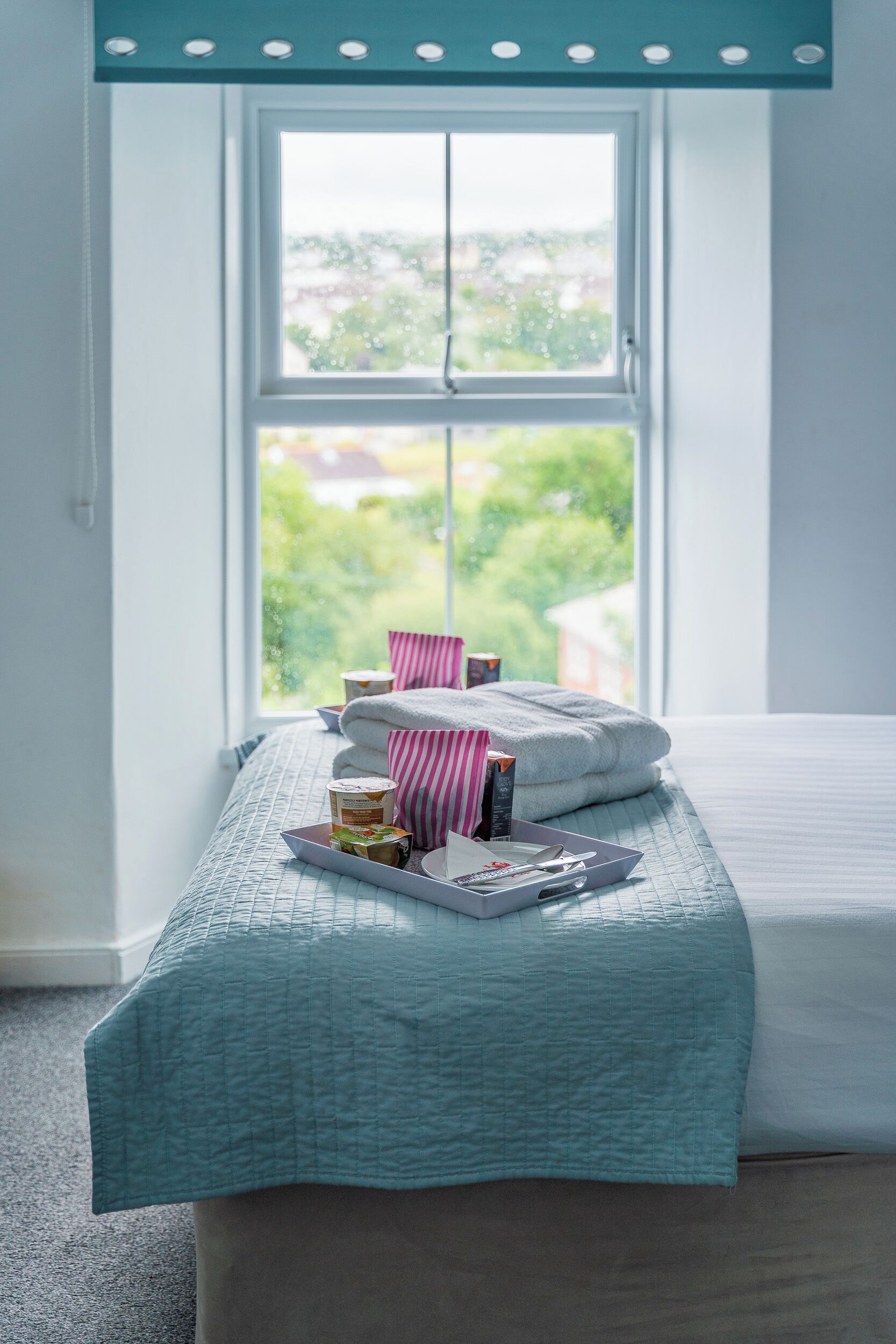 A neatly made bed with a teal quilt, pillows, and a tray with toiletries.