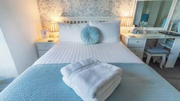 Una cama bien hecha con colcha azul, almohada azul y toalla blanca doblada. Hay una mesita de noche con una lámpara, un espejo y una pequeña silla. La habitación tiene papel tapiz con motivos florales y un cabecero blanco.