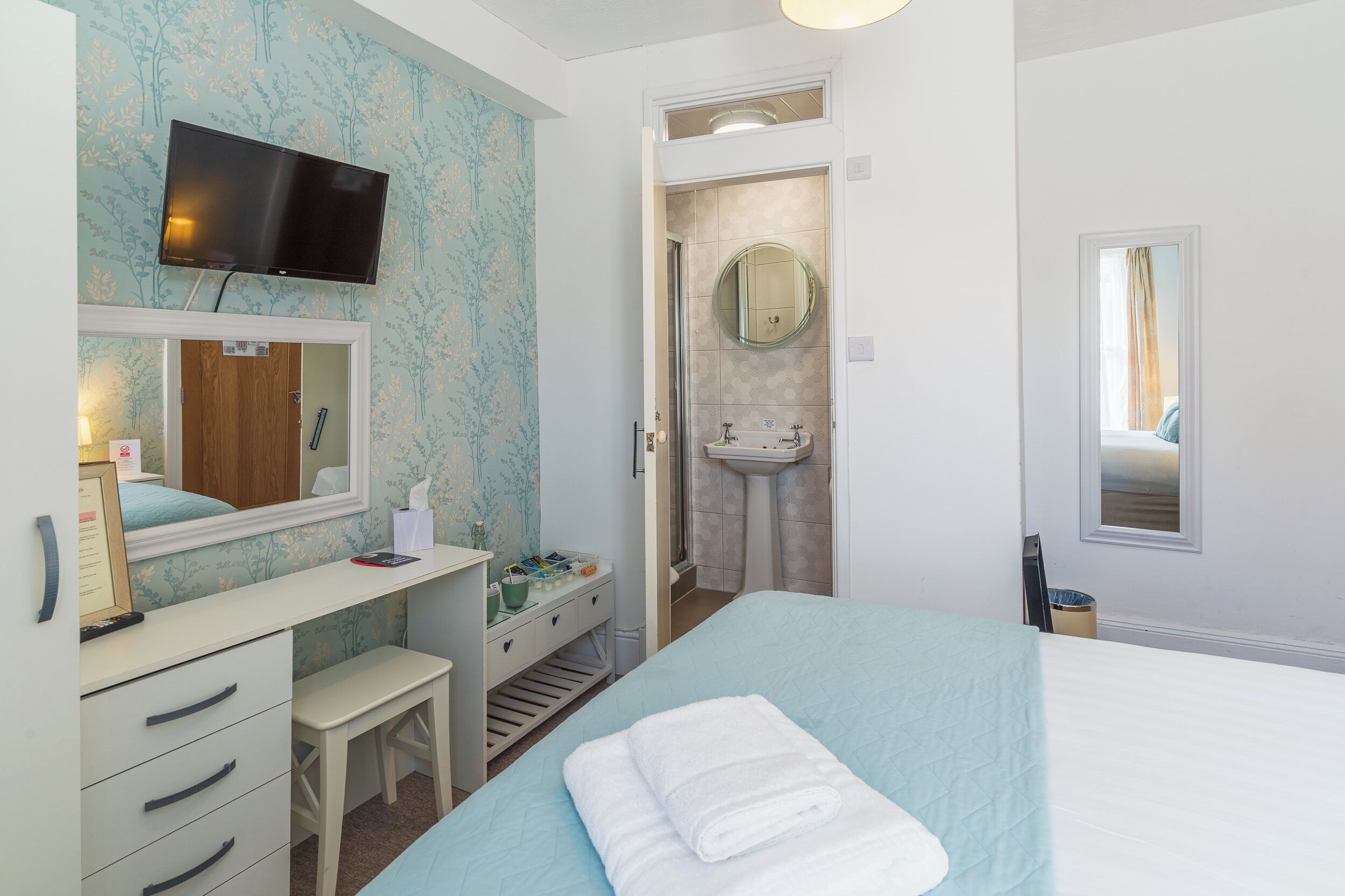 Habitación de hotel con cama, escritorio con cajones, espejo, televisor montado en la pared, baño con lavabo y espejo, y ventana con cortinas.