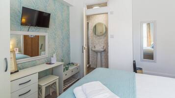 Habitación de hotel con cama, escritorio con cajones, espejo, televisor montado en la pared, baño con lavabo y espejo, y ventana con cortinas.