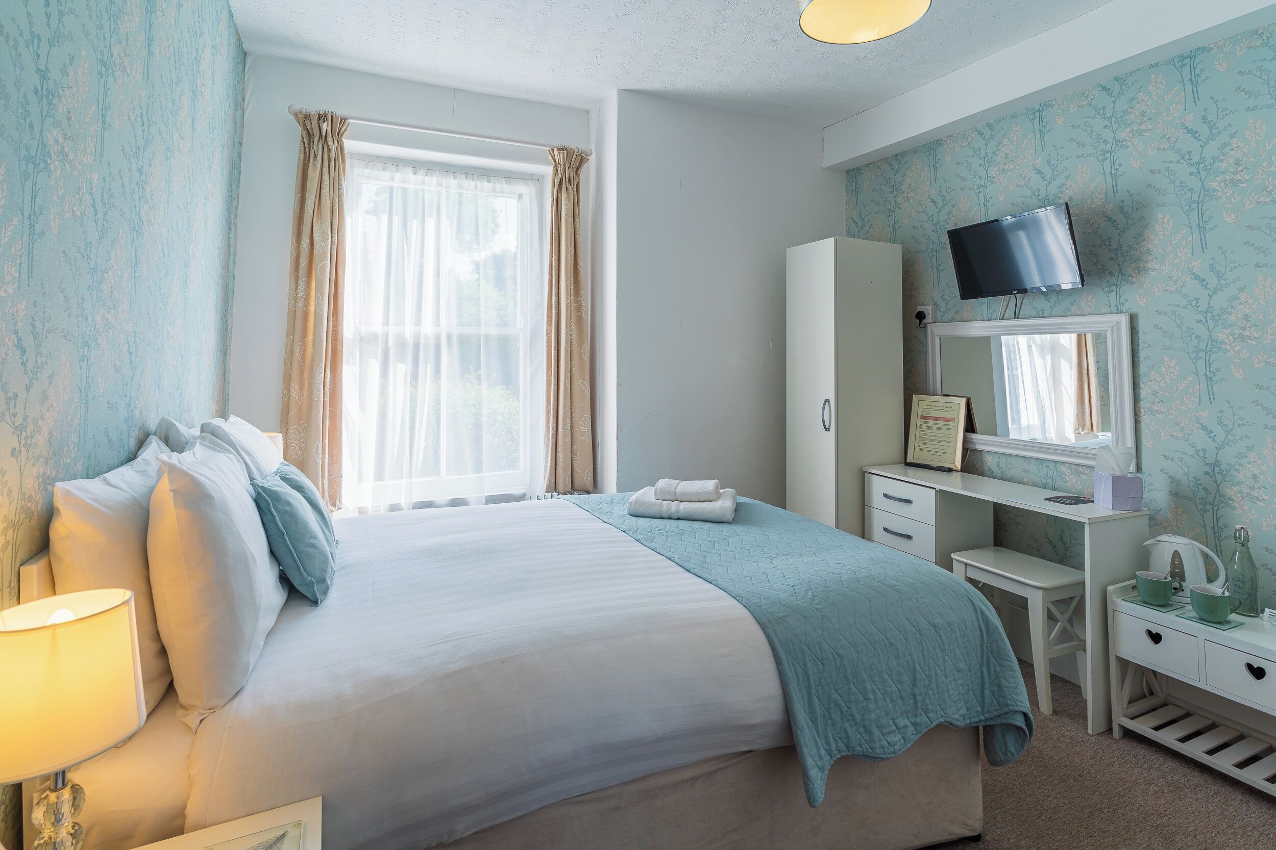 Un dormitorio con cama, escritorio, televisión y ventana con cortinas.