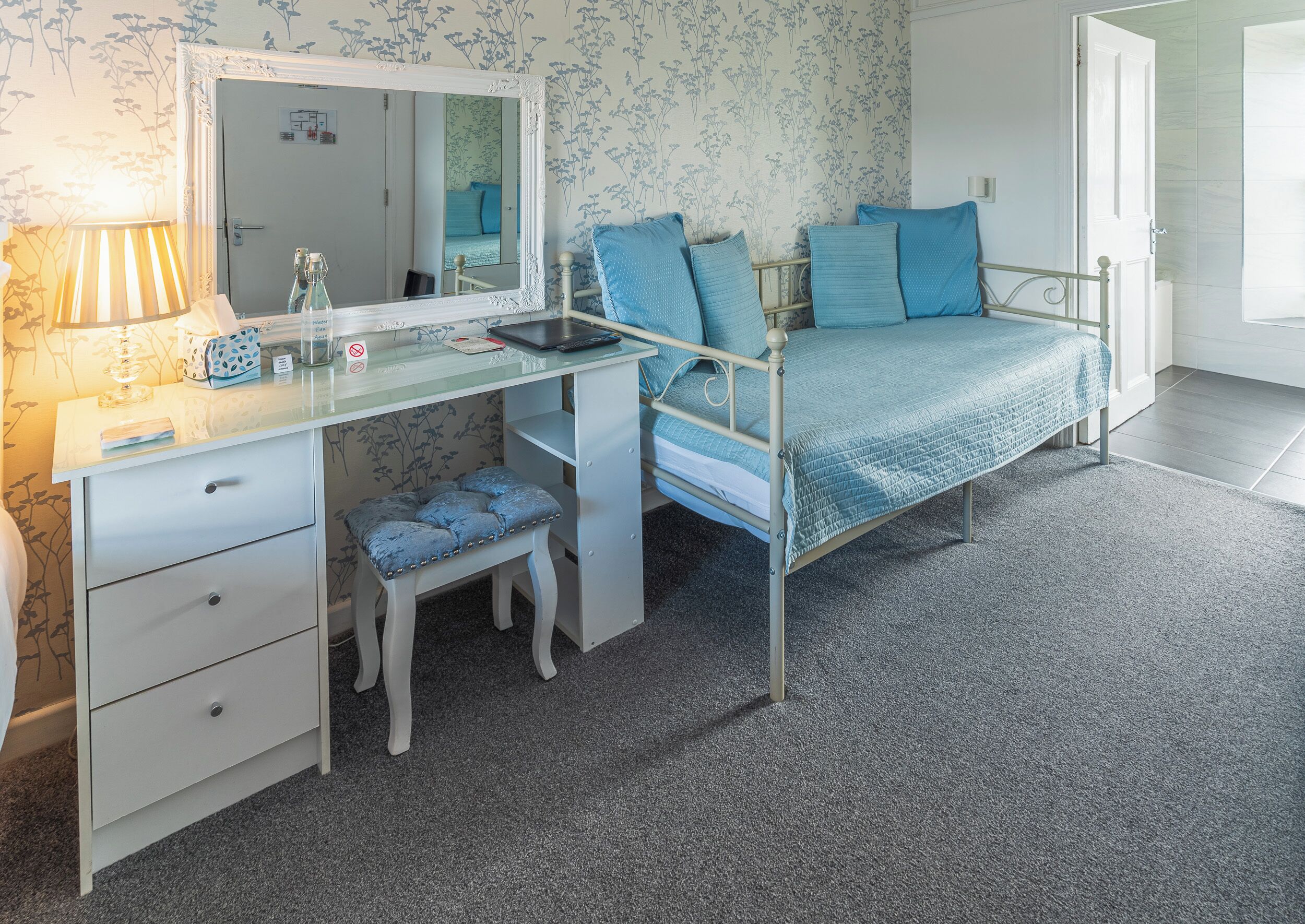 A bedroom with a bed, a desk, a chair, a stool, a mirror, and a lamp.