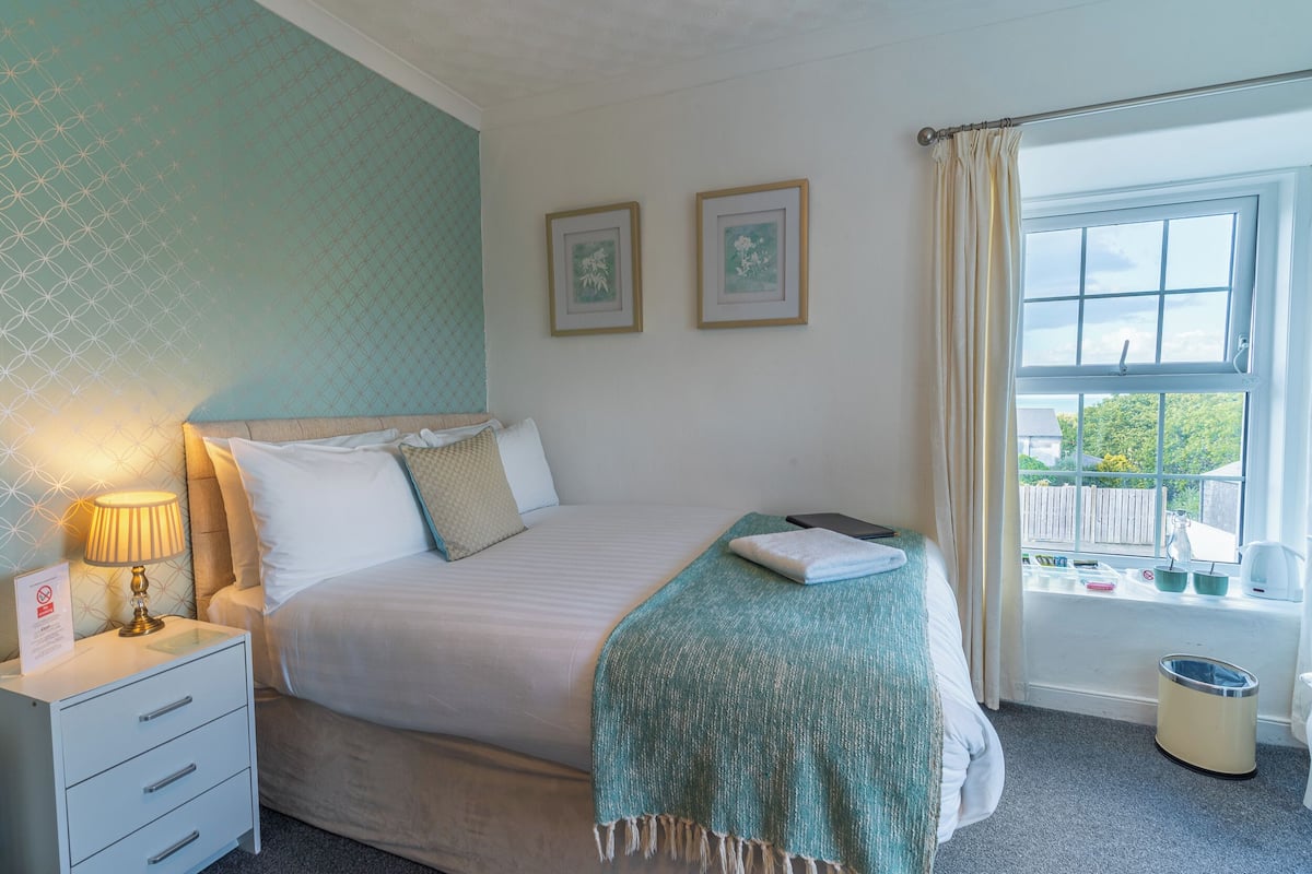 A neatly made bed with a white comforter and a teal throw blanket, a bedside lamp, and a small white nightstand with drawers.