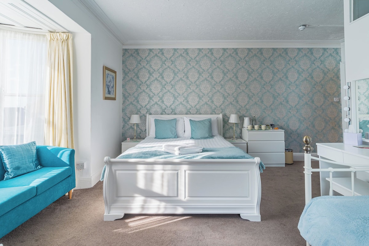 A bedroom with a bed, a nightstand, a dresser, a desk, and a window with curtains.