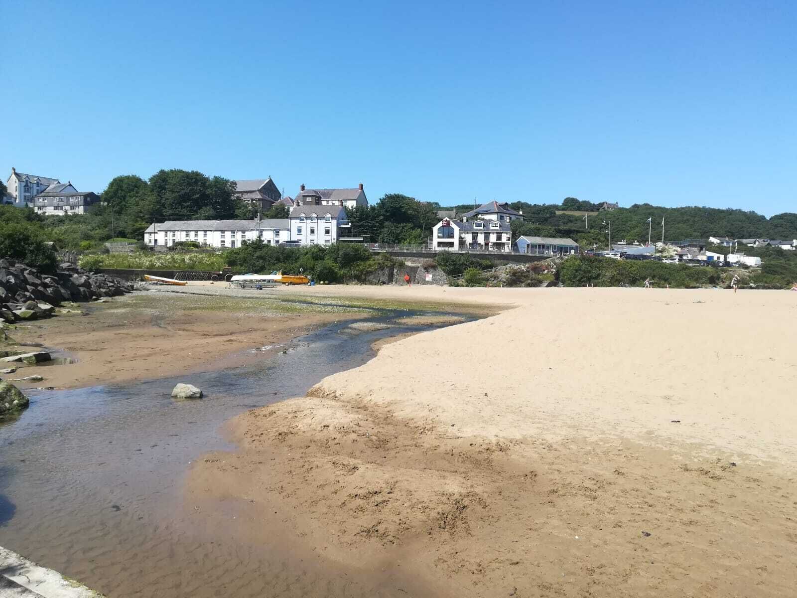Una playa con orilla arenosa y un pequeño arroyo. Se observan edificaciones al fondo, incluyendo una casa grande de color blanco y un edificio más pequeño con tejado azul.
