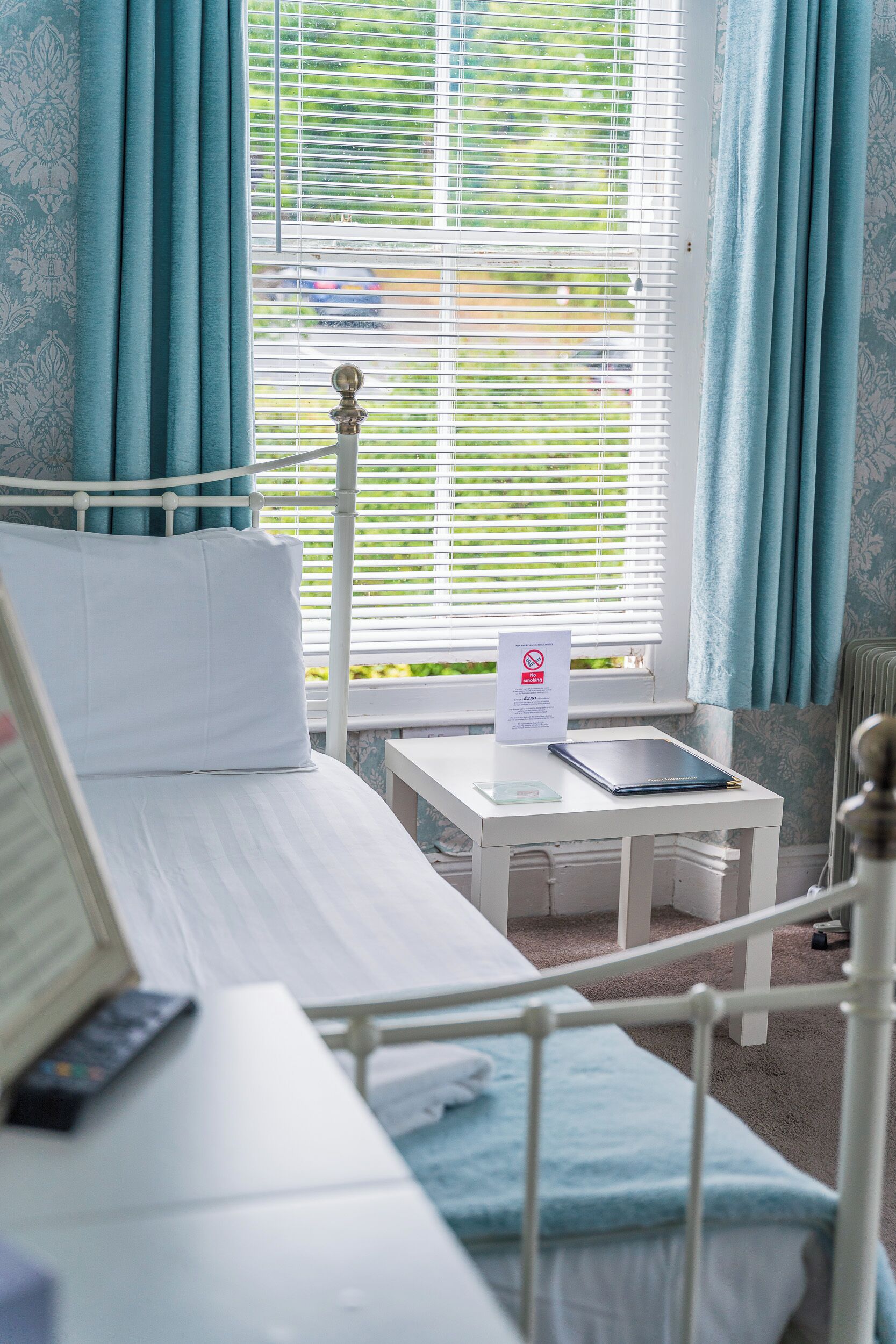 A hotel room with a bed, a bedside table with a remote control, a window with blinds, and a sign on the window.