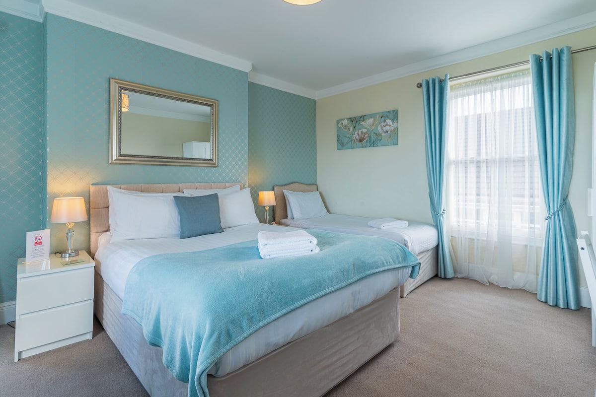 A bedroom with a large bed, two bedside tables, a nightstand, a mirror, and a window with curtains.