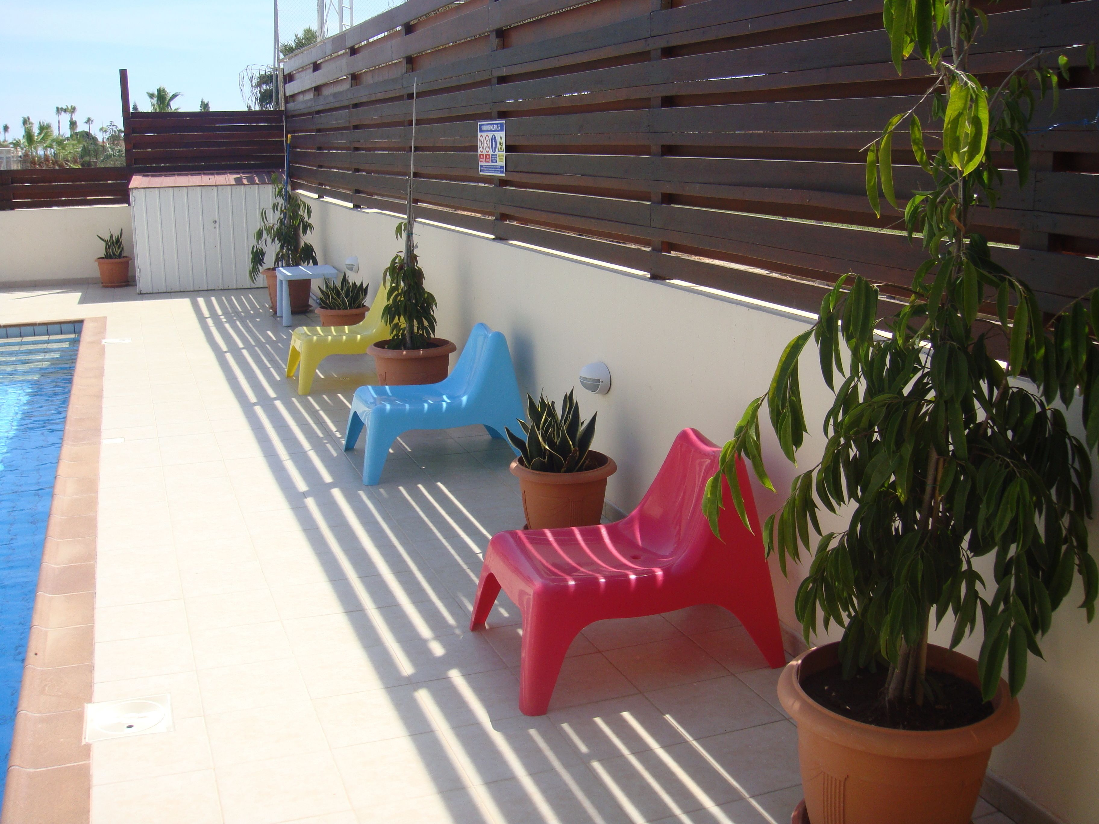 A poolside area with a wooden fence, potted plants, and colorful plastic chairs.