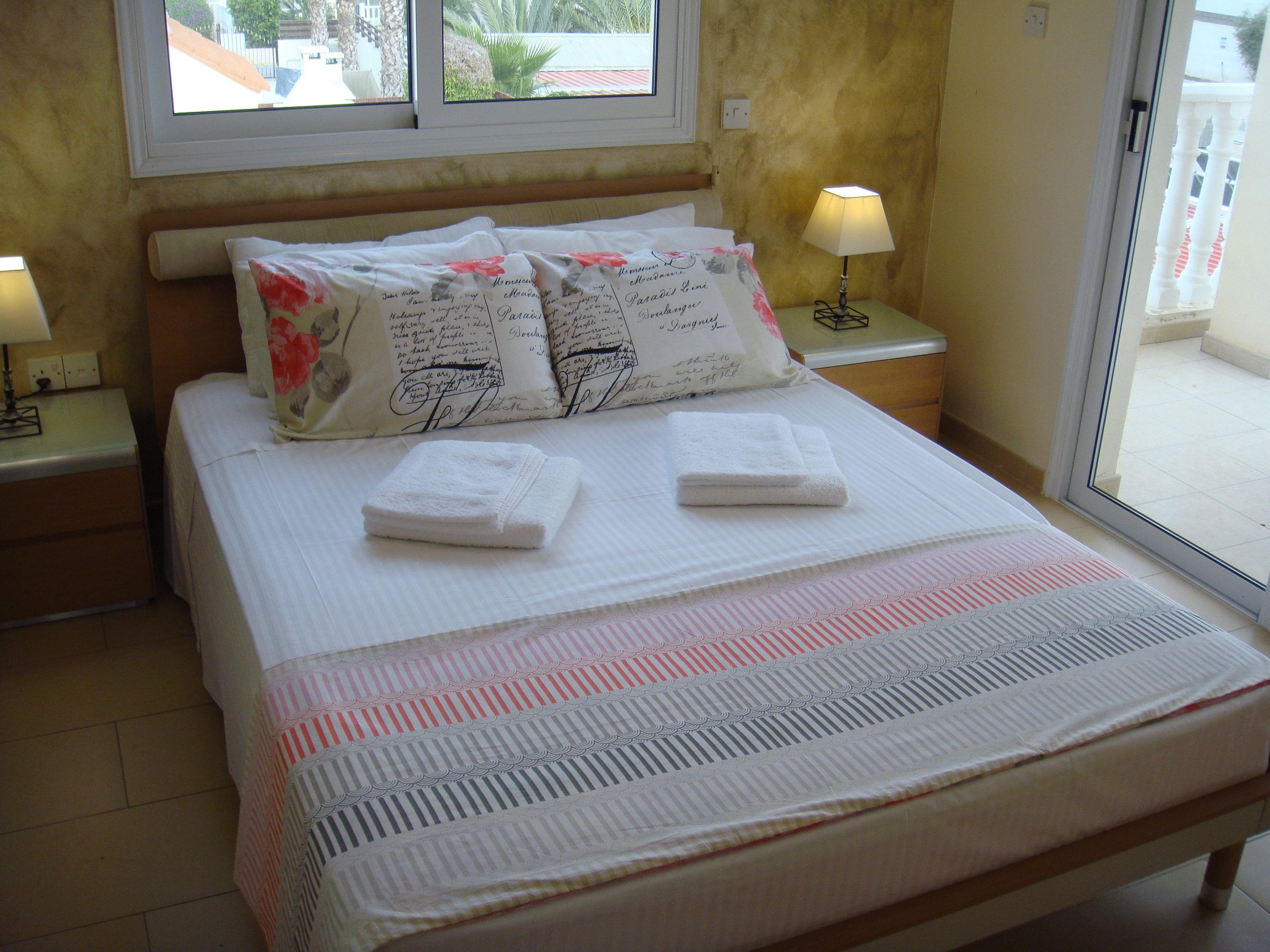A neatly made bed with white linens and two folded towels, a window with a view of the outside, and two bedside lamps on nightstands.