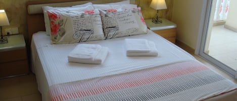 A neatly made bed with white linens and two folded towels, a window with a view of the outside, and two bedside lamps on nightstands.