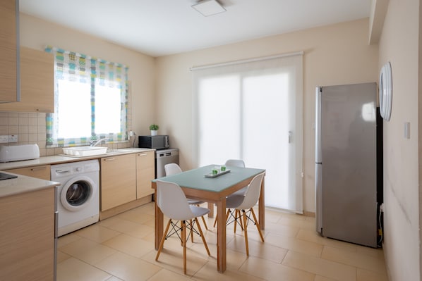 A compact kitchen with a washing machine, a dining table with chairs, a refrigerator, and a window with curtains.