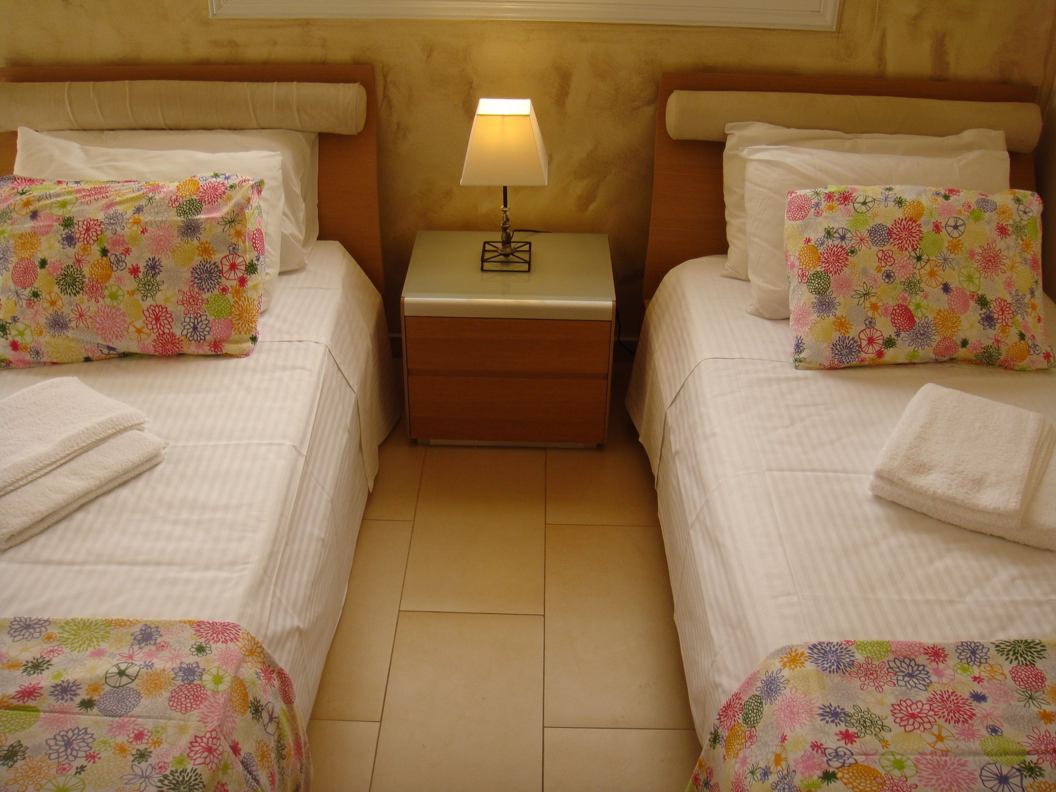 A hotel room with two beds, each with a floral-patterned bedspread and matching pillows. There is a nightstand with a lamp between the beds.