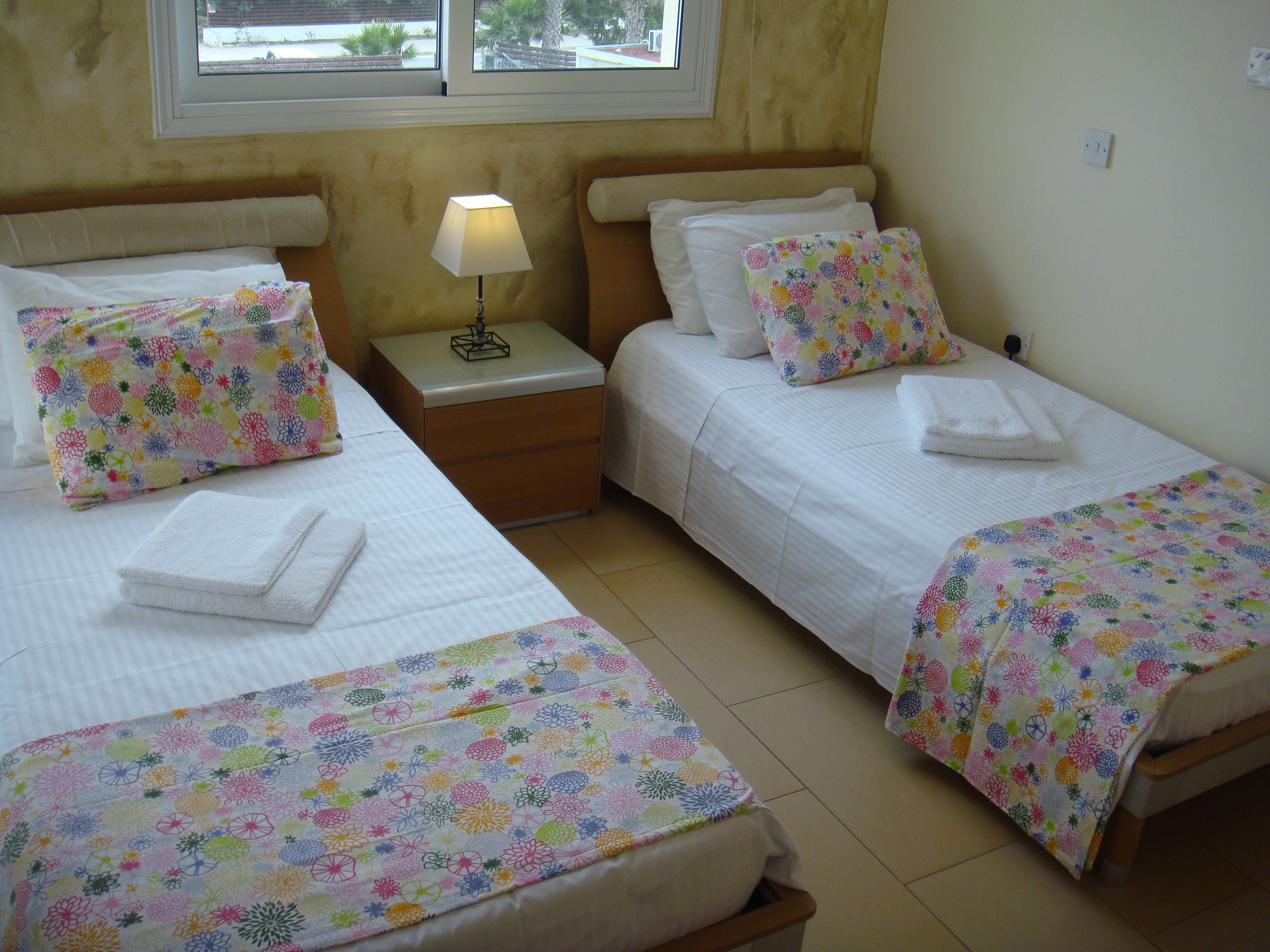 A hotel room with two beds, each with a floral-patterned bedspread and matching pillows. There is a nightstand with a lamp between the beds, and a window above the beds.