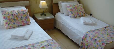 A hotel room with two beds, each with a floral-patterned bedspread and matching pillows. There is a nightstand with a lamp between the beds, and a window above the beds.