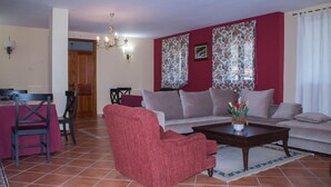 A living room with a red armchair, a sofa, and a coffee table. There is a dining table with chairs and a chandelier. - CR La Flor del Naranjo (Cadalso)