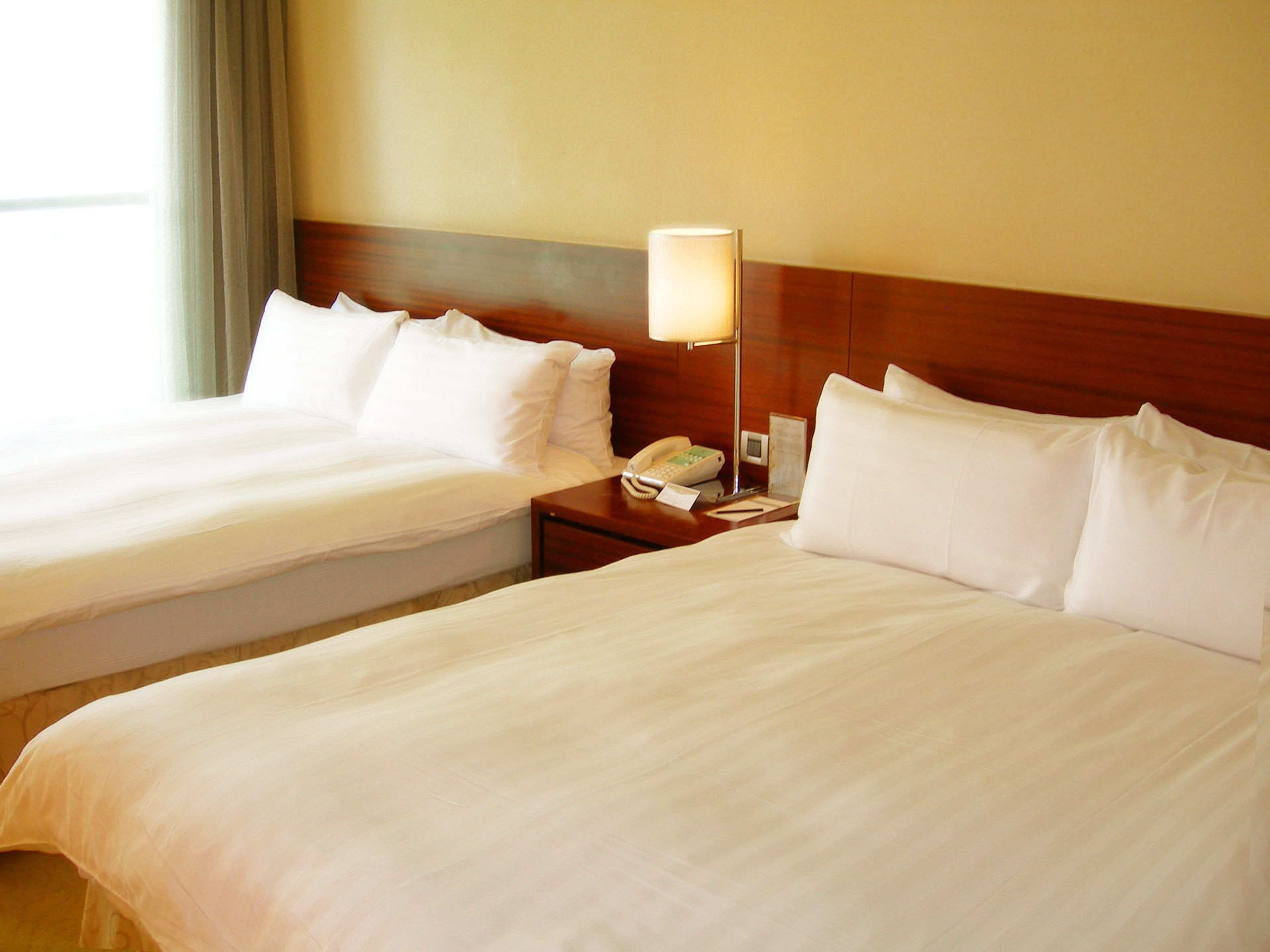 A hotel room with two beds, a bedside table with a lamp, a phone, and a small clock.
