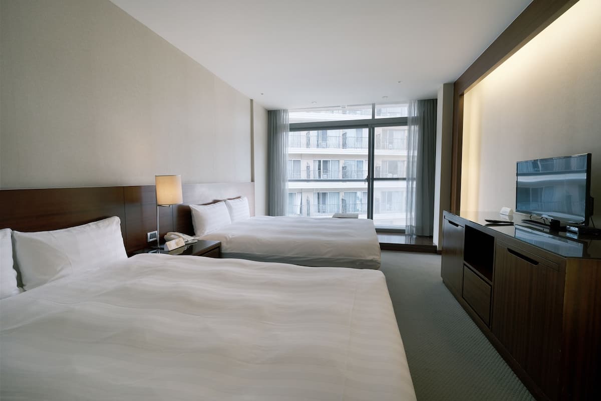 A hotel room with two beds, a TV, a desk, and a large window with a city view.