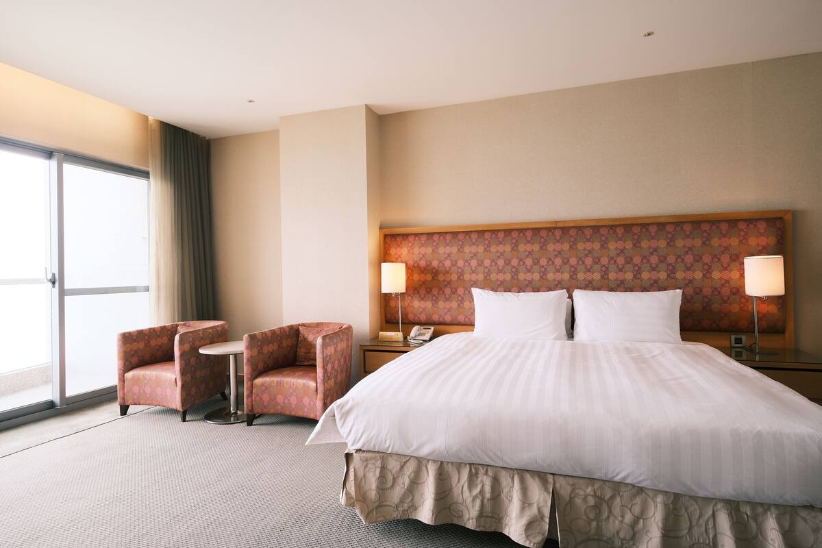 A hotel room with a large bed, two armchairs, a small table, a nightstand with a lamp, and a large window with curtains.