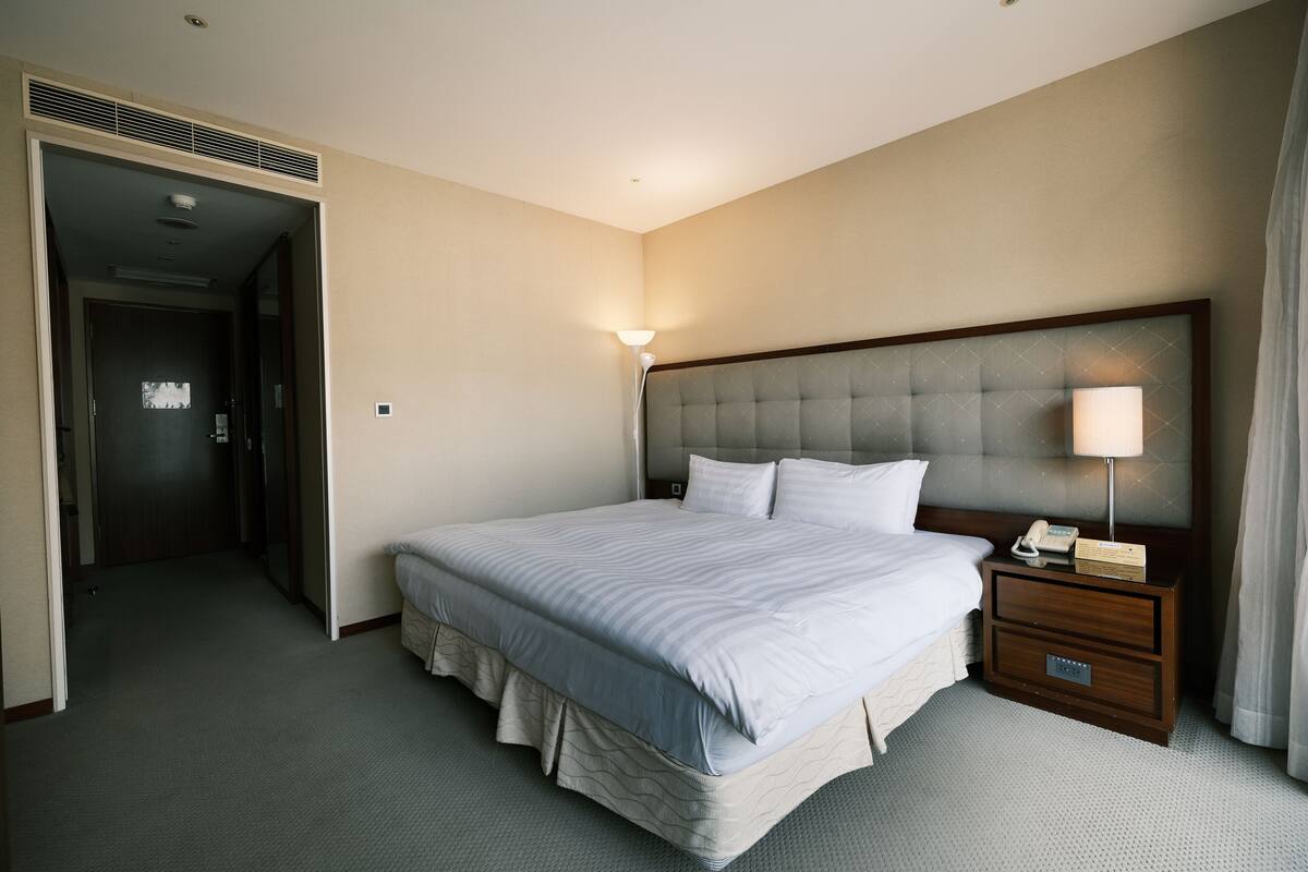 A hotel room with a large bed, a bedside table with a lamp, and a door leading to a hallway.