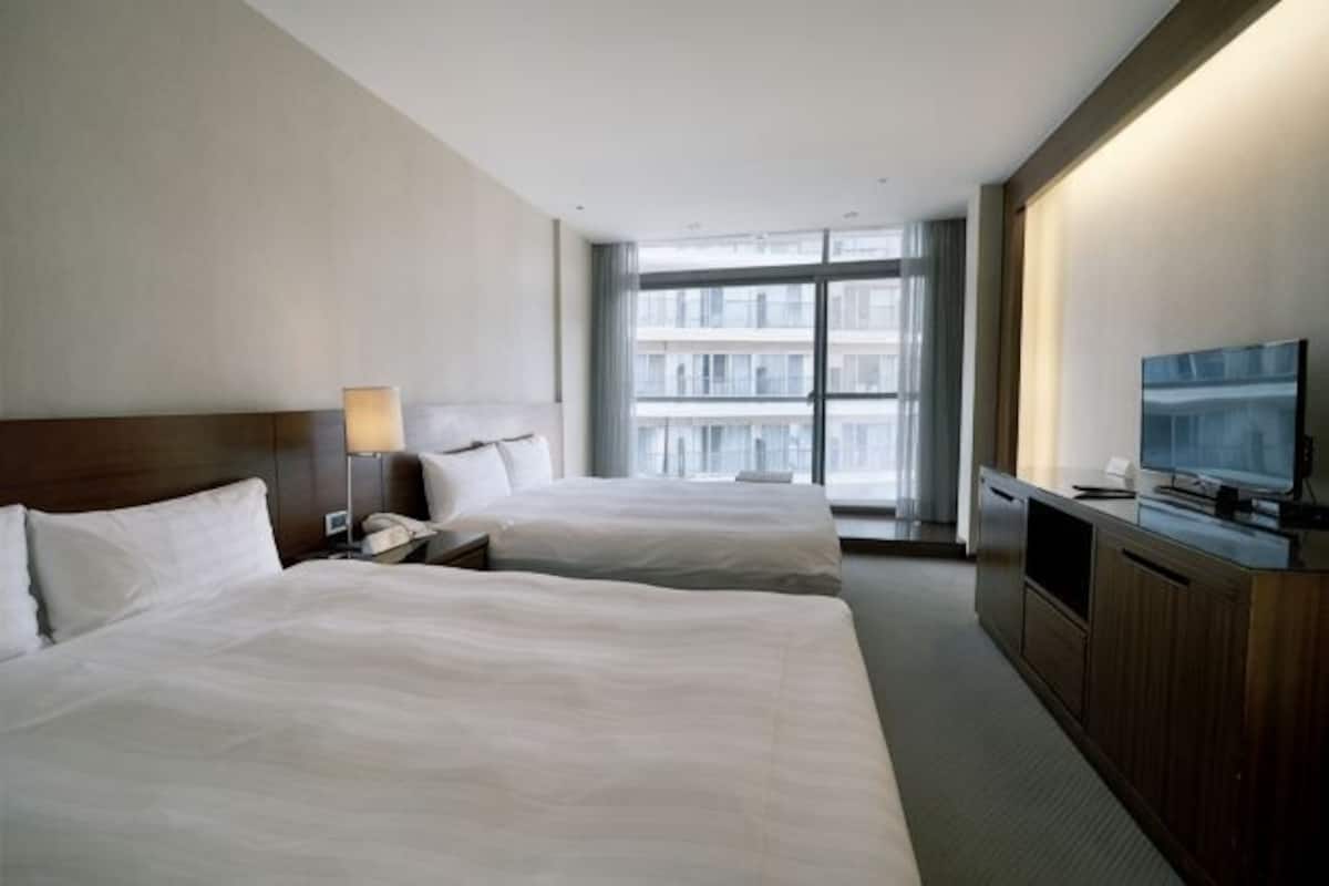 A hotel room with two beds, a TV, a desk, and a large window with a city view.