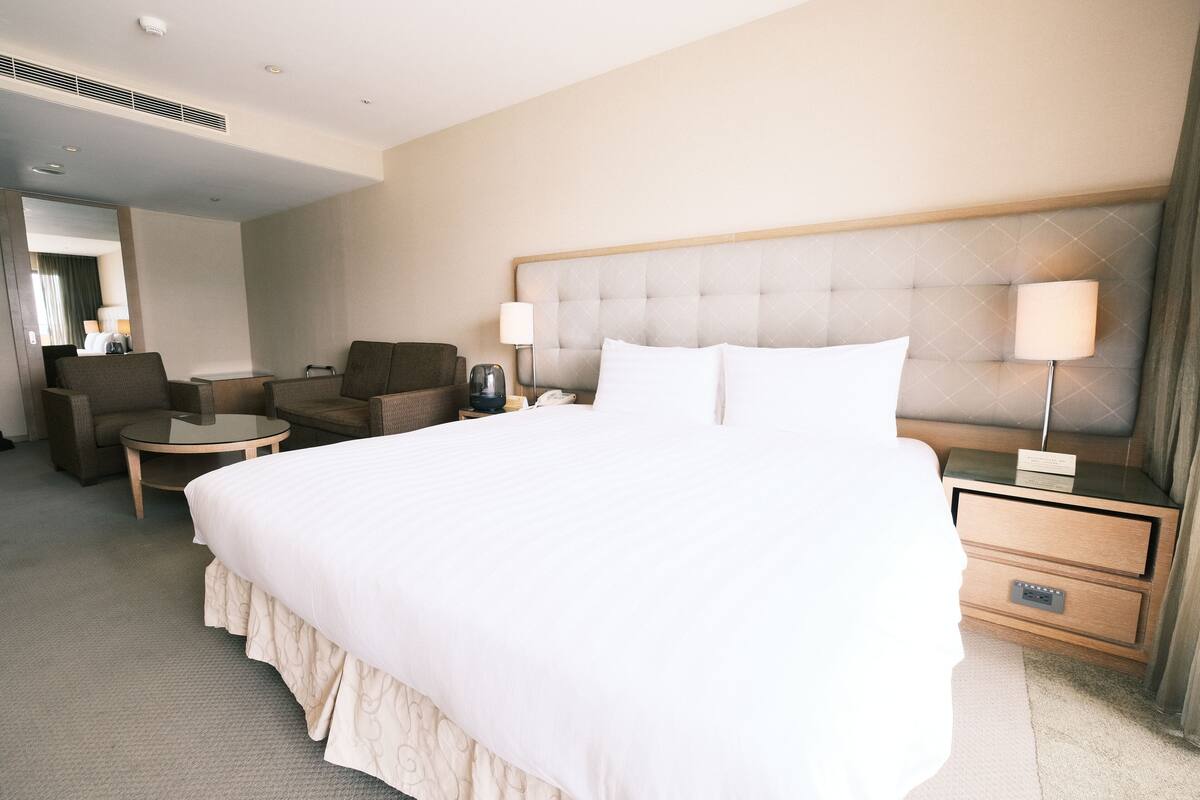 A hotel room with a large bed, a bedside table with a lamp, a chair, and a small table.