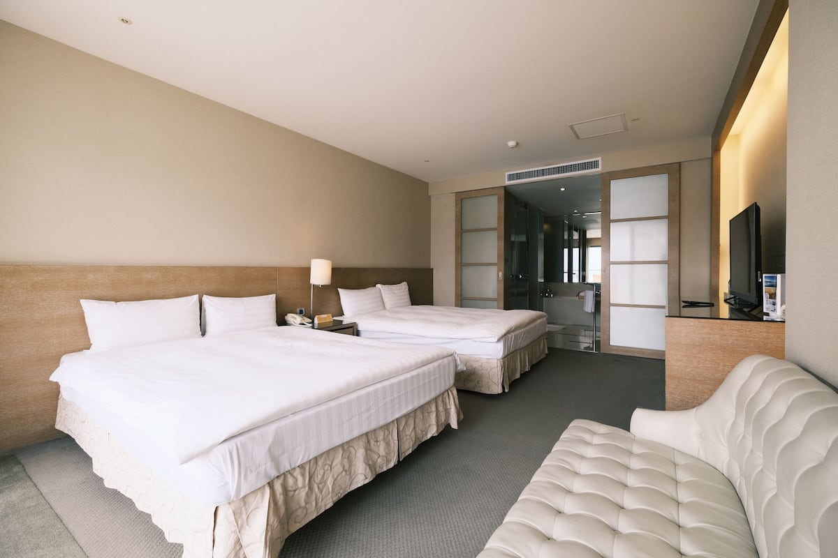 A hotel room with two beds, a sofa, a TV, and a bathroom visible through a glass door.