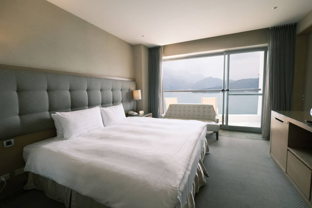 A hotel room with a large bed, a desk, a chair, and a view of the water and mountains.