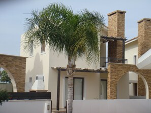 Un edificio de dos pisos con fachada de piedra, un balcón con barandilla metálica y una palmera en primer plano.