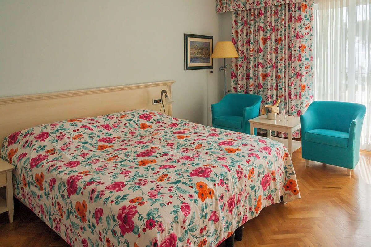 A bedroom with a floral bedspread, two chairs, a nightstand, a lamp, and a painting on the wall.