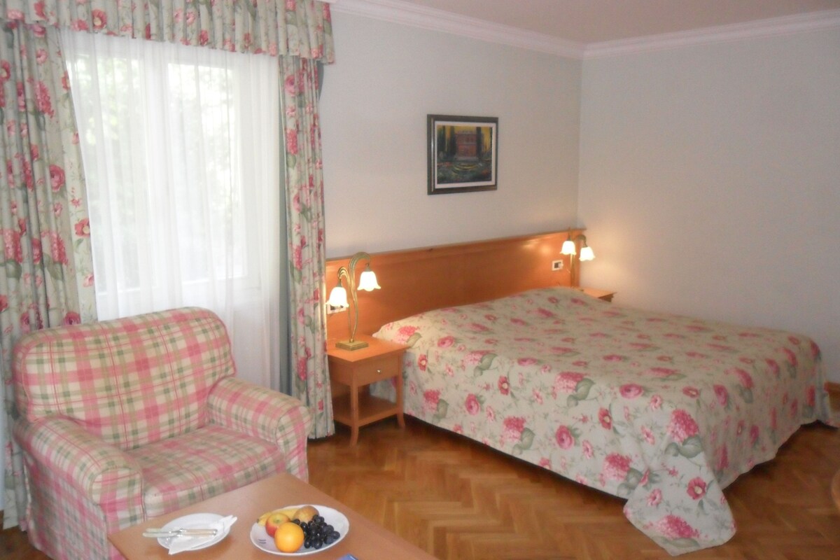 A hotel room with a bed, a sofa, a bedside table with a lamp, and a plate of fruit on a small table.
