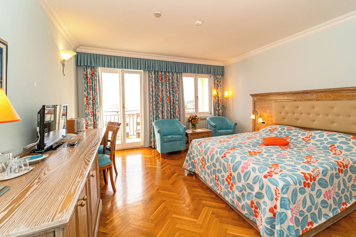 A hotel room with a wooden desk, two armchairs, a bed with a floral bedspread, and a balcony with curtains.