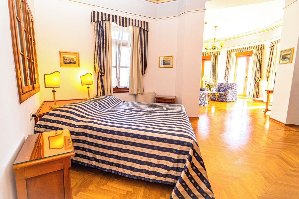 A hotel room with a large bed, two bedside lamps, a window with curtains, a radiator, a small table, and a mirror.