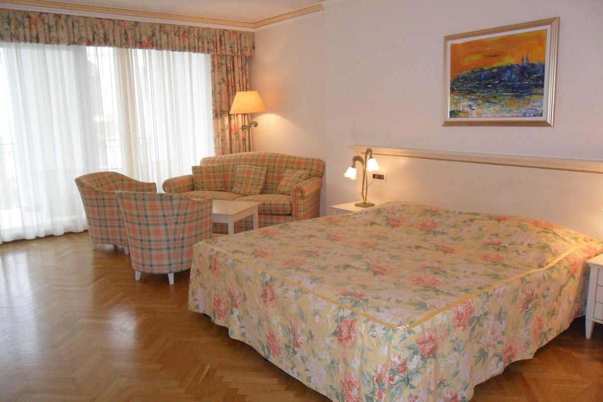 A hotel room with a bed, a sofa, two armchairs, a small table, a lamp, a painting on the wall, and a window with curtains.