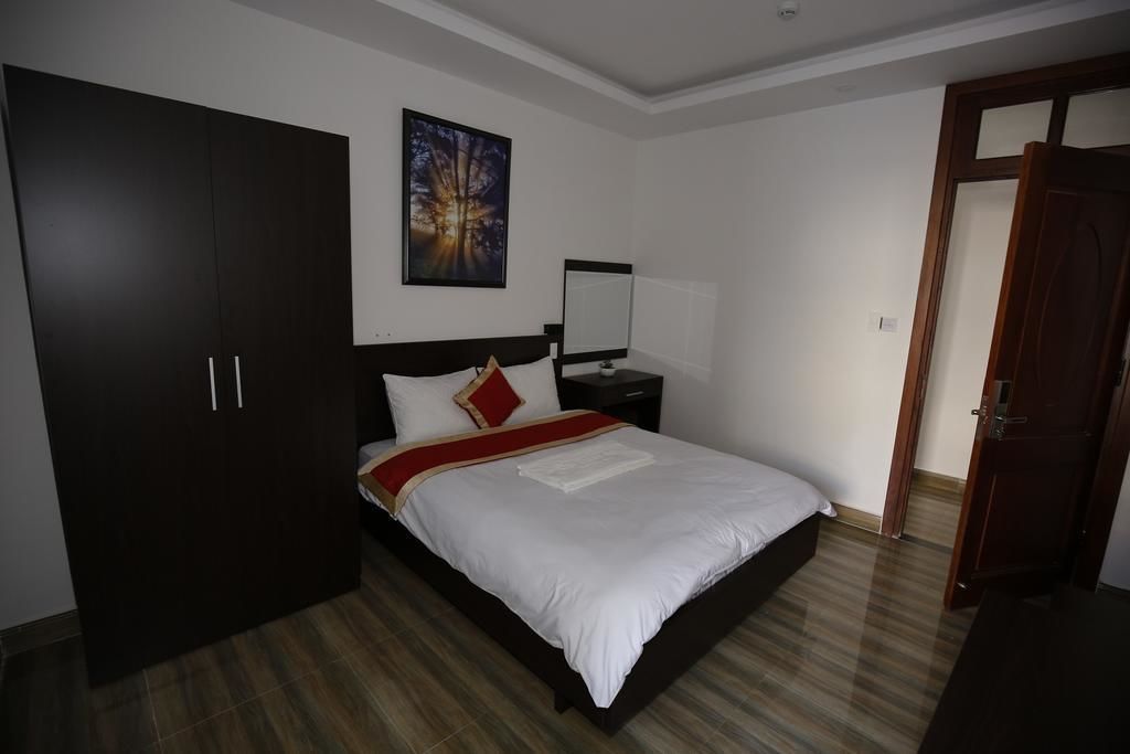 A bedroom with a large dark wardrobe, a bed with white bedding, a red throw, and a nightstand with a lamp.