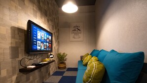 Ein Zimmer mit einem an der Wand befestigten Flachbildfernseher, einem blauen Sofa mit grünen Kissen, einer kleinen Pflanze und einem gerahmten Motivationsspruch.