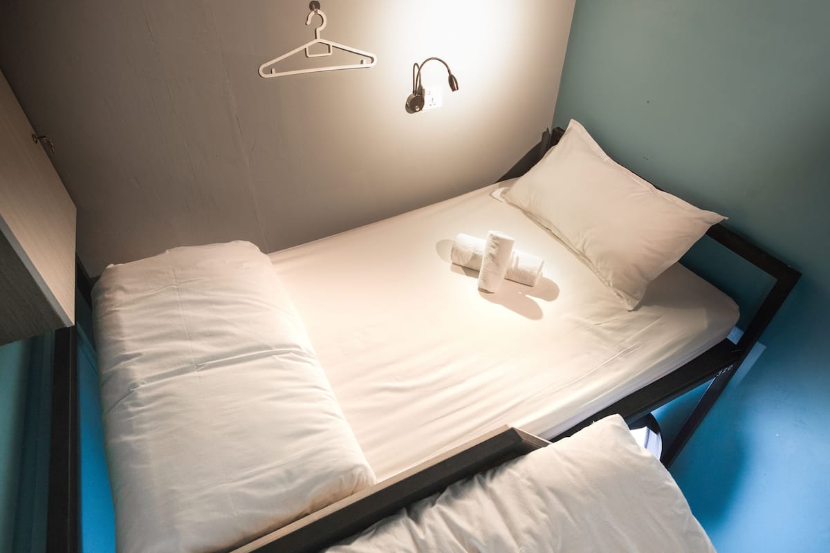 A single bed with white linens, a hanging coat rack, and a wall-mounted light fixture.