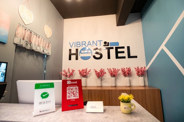 A reception desk with a sign reading "Vibrant Hostel," a QR code for Boost payment, and a small table with a vase of flowers. - Vibrant Hostel (Kota Kinabalu)