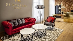 Un hall d’hôtel moderne avec un canapé rouge, une table basse ronde, un tapis à motifs et un comptoir de réception adossé à un mur en pierre.