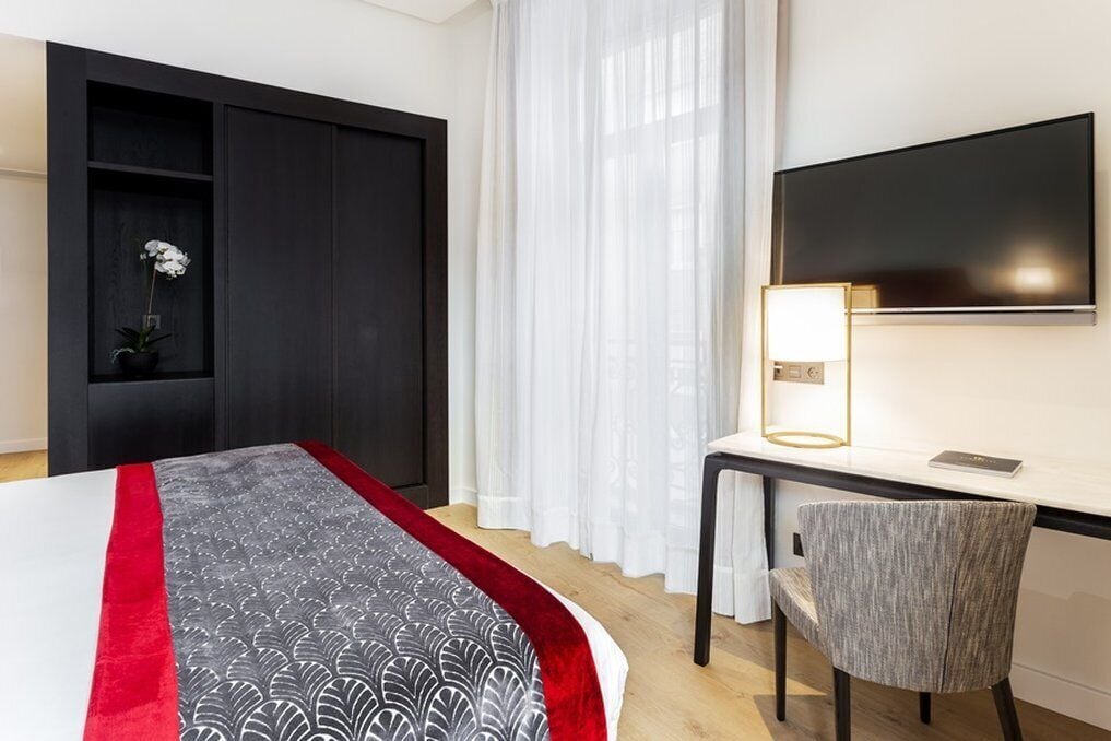 A modern hotel room with a bed, a desk with a lamp, a chair, and a TV.