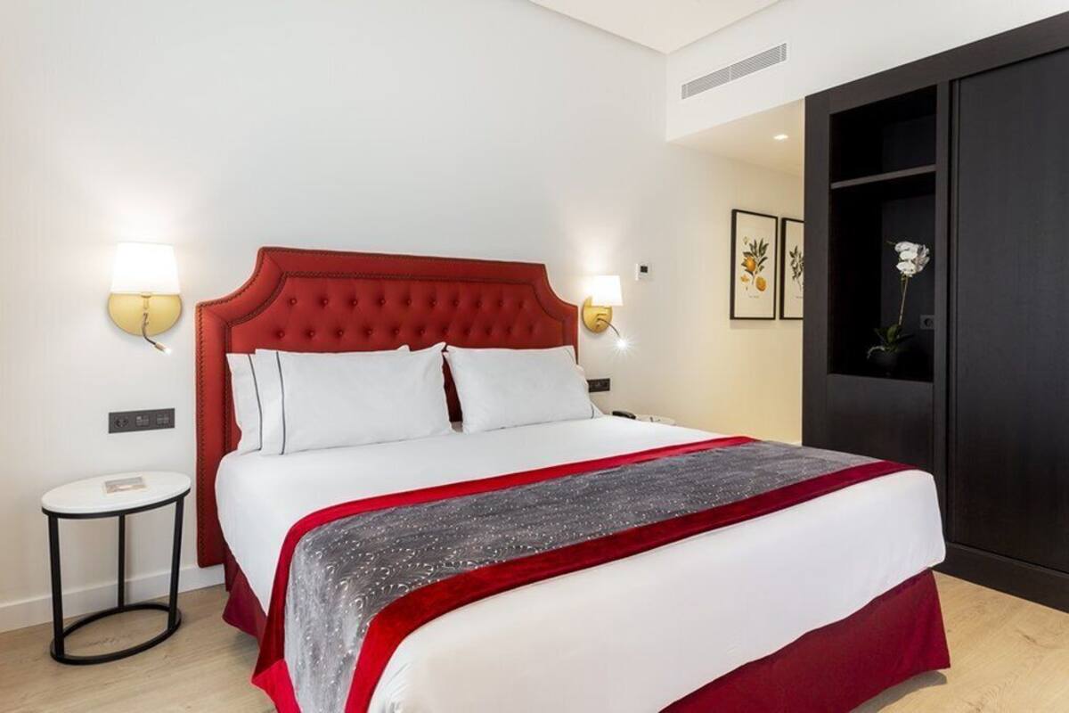 Een netjes opgemaakt bed met een rode hoofdbord, witte beddengoed en een grijze decoratieve plaid. Er staan twee nachtkastjes met lampen, een klein rond tafeltje en een donkere garderobe.