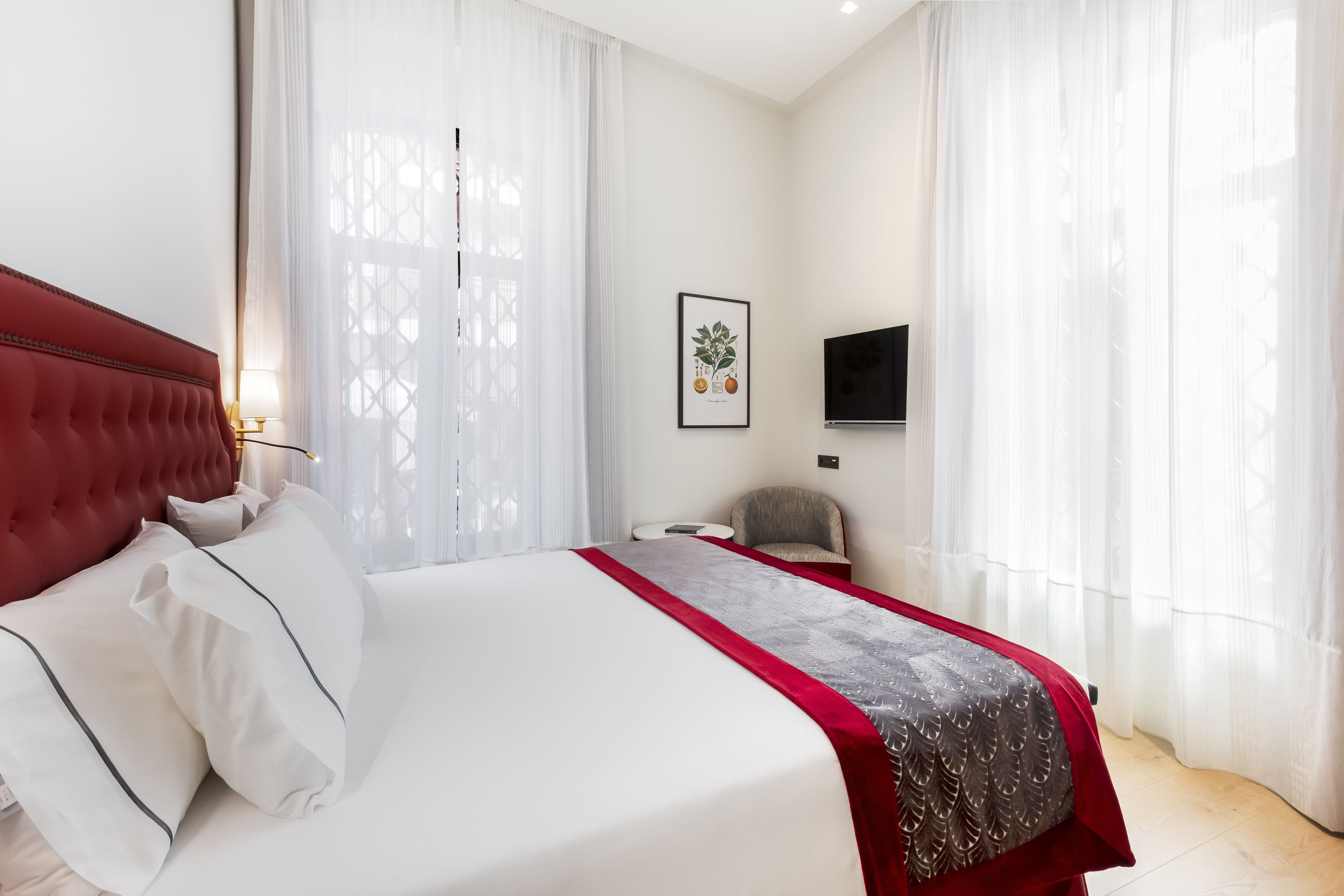 Ein Hotelzimmer mit einem großen Bett, einem roten Kopfteil, weißer Bettwäsche, einer grau-roten Tagesdecke, einem Sessel, einem Fernseher an der Wand und transparenten Vorhängen.