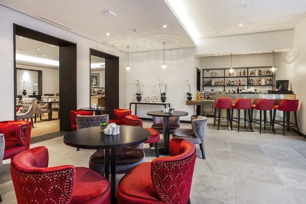 A modern hotel lobby with red upholstered chairs, a bar area with red bar stools, and a seating area with round tables.