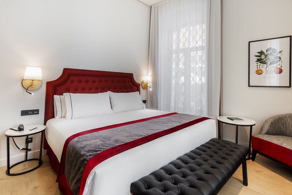 A hotel room with a red upholstered bed, a black tufted bench, a small round table, and a wall-mounted lamp.