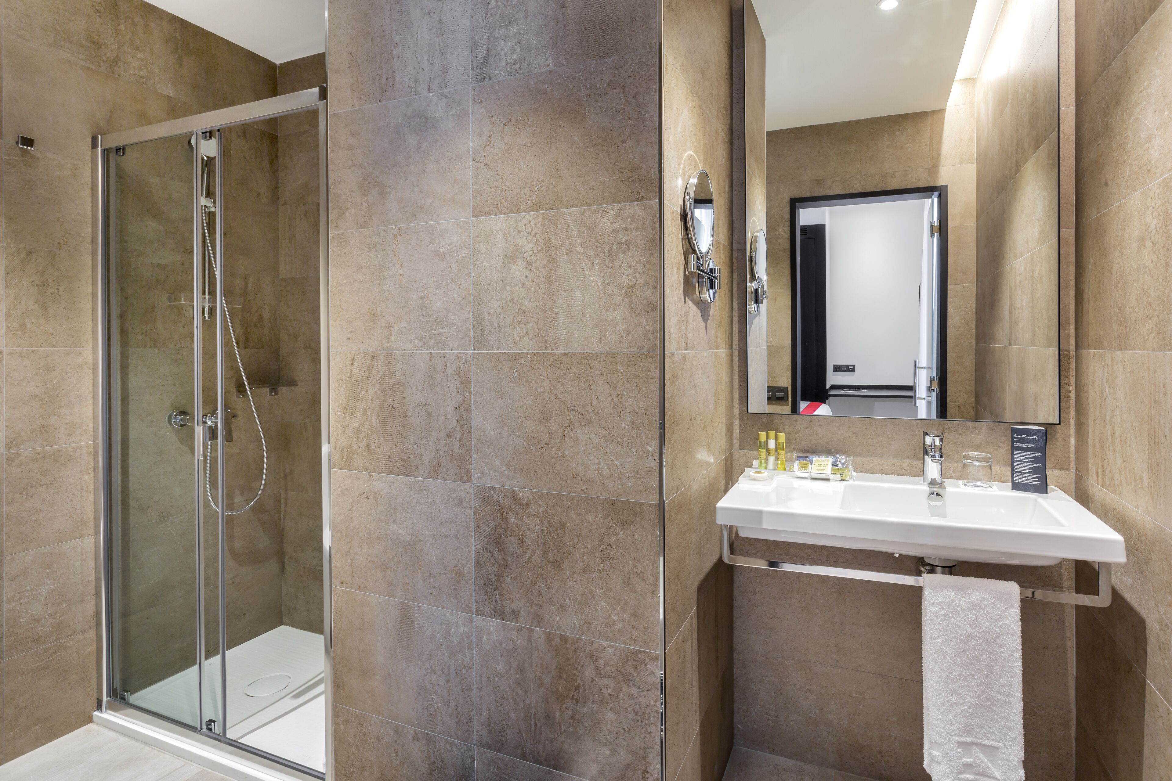 Un baño moderno con ducha envuelta en vidrio, un lavamanos blanco con espejo y azulejos color beige.