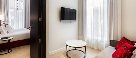 Una habitación de hotel moderna con una cama, una pequeña mesa redonda, un televisor montado en la pared y un sofá con cojines rojos.