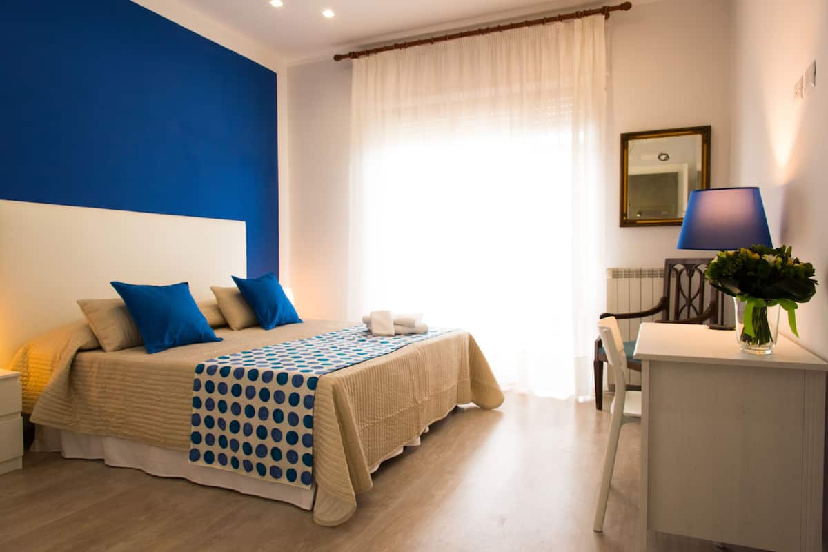 Une chambre à coucher avec un lit, un bureau, une chaise, une lampe et une fenêtre avec des rideaux.