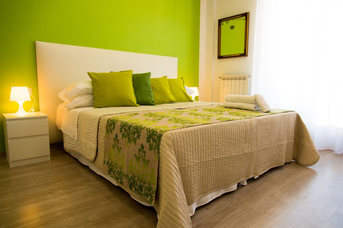 Un lit bien fait, recouvert d’une couvre-lit verte et beige, avec des oreillers blancs et une tête de lit blanche. On y trouve une table de chevet avec une lampe, ainsi qu’une lampe de chevet blanche. La pièce est dotée d’un parquet en bois clair et d’un mur vert.